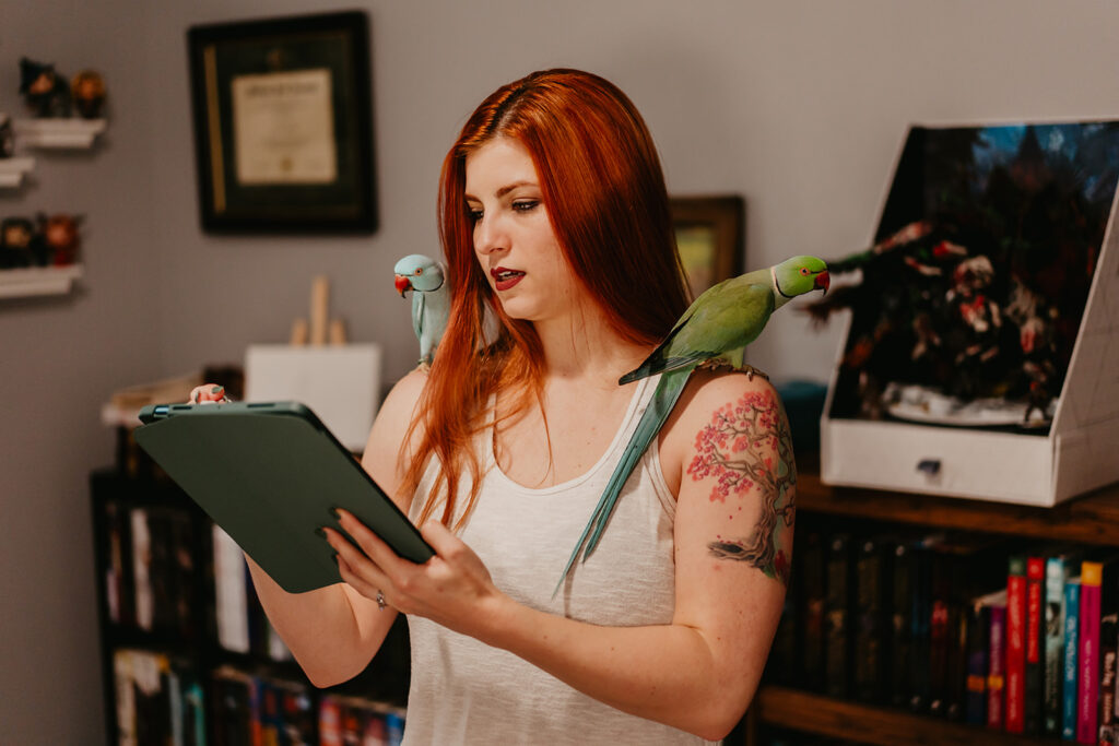A red-haired woman wearing a white tank top that shows off a tree tattoo on her left shoulder writes or draws on an iPad, while two parrots sit on either shoulder. On her right shoulder is a blue parrot with a red beak and on her left is a green-to-blue parrot with a red beak and a long tail.