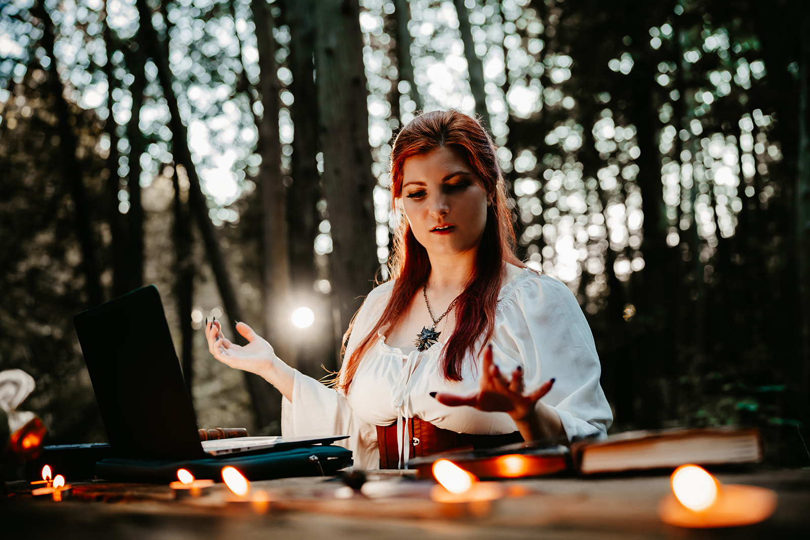 a young woman with red hair, wearing renaissance garb and a witcher medallion sits at a picnic table in the woods with the sun setting behind the trees. She appears to be casting a spell over her computer, tarot cards, and journals, while tea light candles gently burn in the blurred foreground.